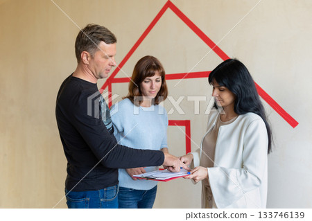 Realtor signs contract, shows plan of apartment in new building to young couple. Concept new homes, new beginning. High quality photo 133746139