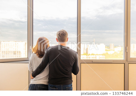 Man hugging his wife. Young couple moving in new apartmen. People looks out window at new houses. Concept new homes, new beginning. High quality photo Man hugging his wife. Young couple moving in new apartmen. People looks out window at new houses. Concept new homes, new beginning. High quality photo 133746244