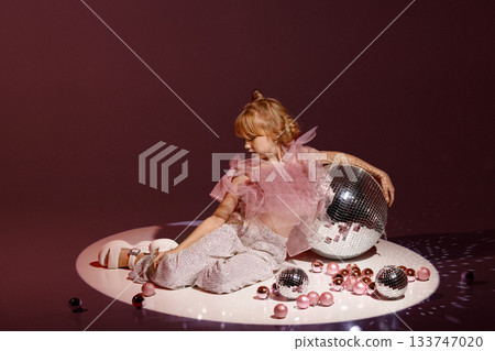 Studio portrait of young girl in festive outfit with disco balls on burgundy background Studio portrait of young girl in festive outfit with disco balls on burgundy background 133747020