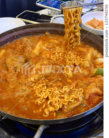 A hearty bowl of Budaejjigae (Korean Army Stew), filled with sausage, tofu, kimchi, and noodles in a spicy broth. This flavorful dish brings warmth and comfort, perfect for sharing. 133748556