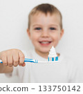 A smiling young boy holds a toothbrush with a swirl of white toothpaste in shape of a Christmas tree. The toothbrush features blue and white colors 133750142