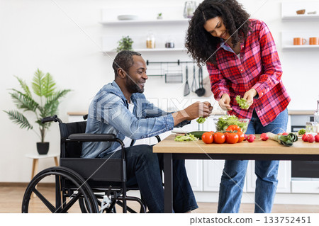An African American couple prepares a meal together in a bright, modern kitchen. One person is in a wheelchair. An African American couple prepares a meal together in a bright, modern kitchen. One person is in a wheelchair. 133752451