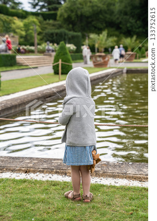 A child in a hooded sweater stands still by the pond, watching the ripples move in soft afternoon light. A child in a hooded sweater stands still by the pond, watching the ripples move in soft afternoon light. 133752615