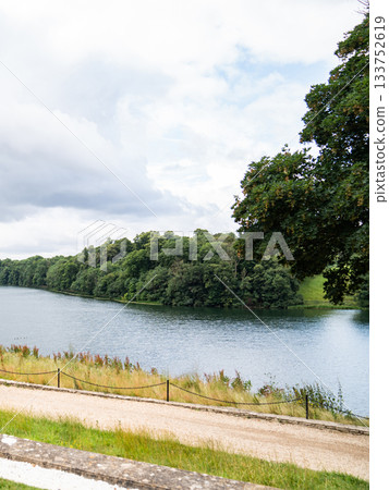 Calm water stretches between green trees under a grey sky, with a small wooden house at the edge. Calm water stretches between green trees under a grey sky, with a small wooden house at the edge. 133752619