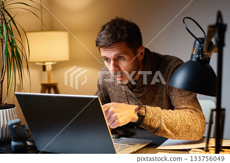 Surprised man looking at laptop screen while working at home office 133756049