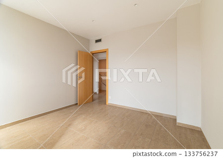 Empty room with beige walls and wooden door. The floor is tiled in light brown. Natural light enters through the open door, creating a spacious feel. 133756237