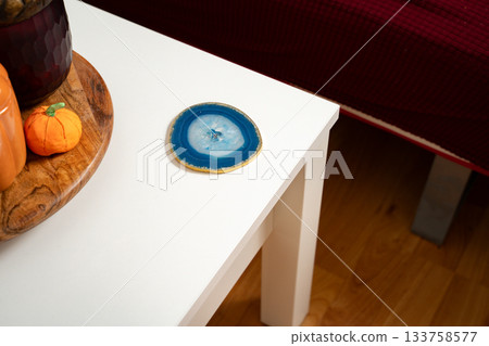 Blue agate coaster placed on the side of a white coffee table in a simple everyday home setting, mixing natural stone with clean modern decor. Concept of energy, grounding, spirituality, minimalism. Blue agate coaster placed on the side of a white coffee table in a simple everyday home setting, mixing natural stone with clean modern decor. Concept of energy, grounding, spirituality, minimalism. 133758577