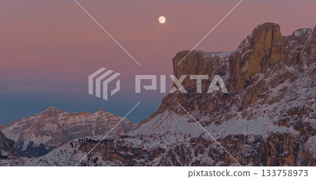 Panoramic view of Dolomite mountain peaks under a glowing full moon. Early winter landscape with snow covered summits in soft dawn light. Panoramic view of Dolomite mountain peaks under a glowing full moon. Early winter landscape with snow covered summits in soft dawn light. 133758973