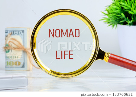 A magnifying glass focuses on the words nomad life, symbolizing freedom and adventure. Nearby, a bundle of dollars and a potted plant add charm to the serene workspace 133760631