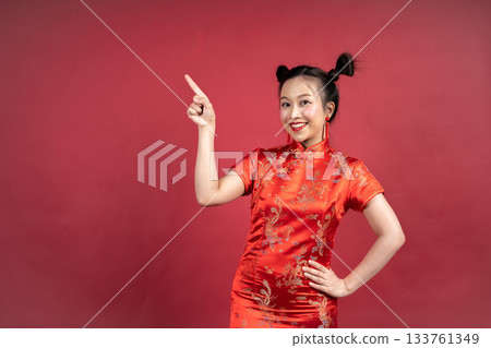 Happy Asian woman in Cheongsam pointing finger at copy space on red background. Happy Asian woman in Cheongsam pointing finger at copy space on red background. 133761349