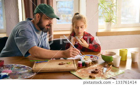 Father guiding daughter while painting clay figures at bright craft table 133761414