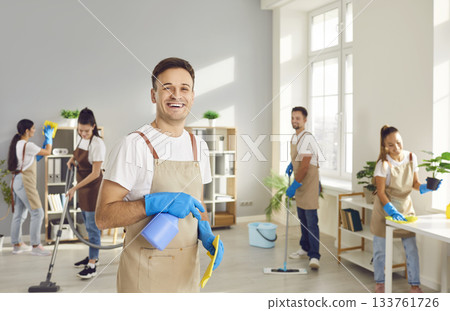 Young janitor looking at camera with a team of cleaning service washing office in background. Young janitor looking at camera with a team of cleaning service washing office in background. 133761726