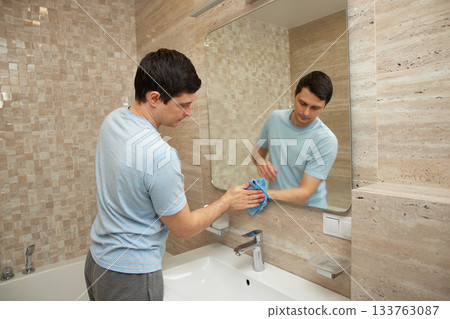 Man cleaning a bathroom mirror reflecting in the glass, doing household chores Man cleaning a bathroom mirror reflecting in the glass, doing household chores 133763087