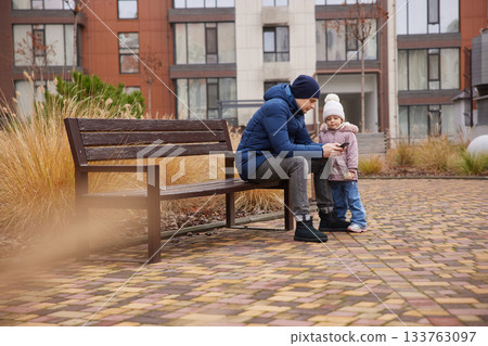 Father and child engaging with a smartphone while sitting on a bench in an urban setting Father and child engaging with a smartphone while sitting on a bench in an urban setting 133763097