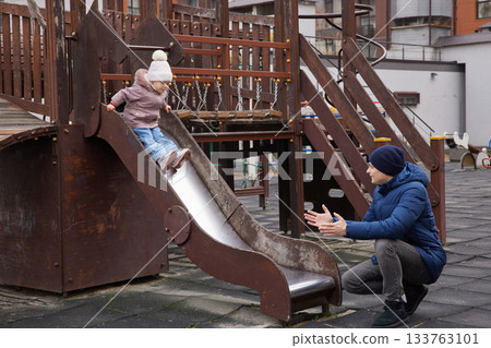 Father catching his toddler daughter sliding down a playground slide 133763101