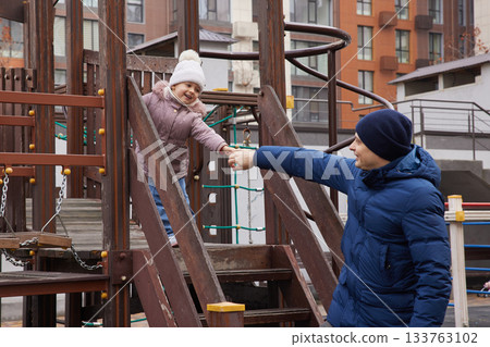 Caring dad assisting child enjoying outdoor play on urban playground in winter Caring dad assisting child enjoying outdoor play on urban playground in winter 133763102