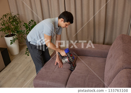 Man vacuuming sofa in living room, performing housework for maintaining home cleanliness 133763108