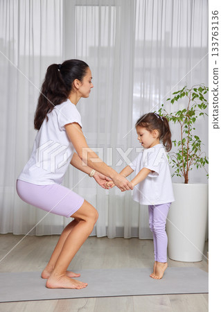 Mother and daughter exercising together on a yoga mat, doing a squat 133763136
