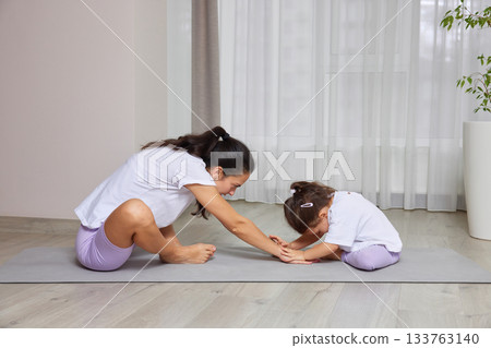 Mother helping young daughter stretching on a yoga mat during home workout 133763140