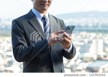 A middle-aged male businessman in a suit holding a smartphone with a town in the background A middle-aged male businessman in a suit holding a smartphone with a town in the background 133763343
