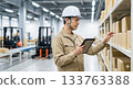 一名戴著安全帽的男工人在物流倉庫操作平板電腦，管理庫存和檢查貨物。 133763388