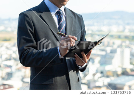 A middle-aged male businessman wearing a suit with a notebook against the backdrop of a city 133763756