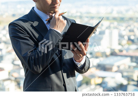 A middle-aged male businessman in a suit holding a notebook and worrying with a city in the background A middle-aged male businessman in a suit holding a notebook and worrying with a city in the background 133763758