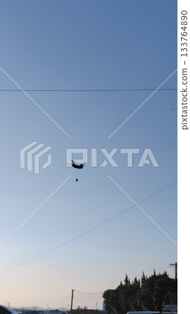 Silhouette of a large helicopter flying with a fire bucket hanging from it 133764890