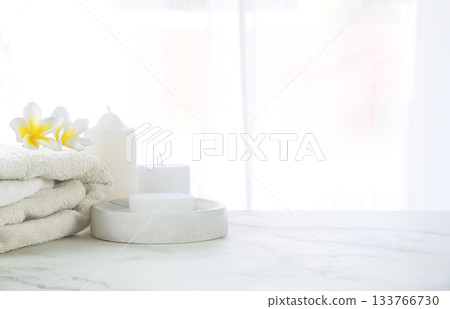 Minimal style soap holder and towel on a luxury stone marble tabletop in a bright bathroom. Decorated with potted plants, flowers and candle, with a blurred curtain of a window background 133766730