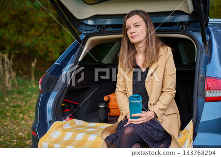 Woman holding thermos cup while sitting on open car trunk with blanket Woman holding thermos cup while sitting on open car trunk with blanket 133768204