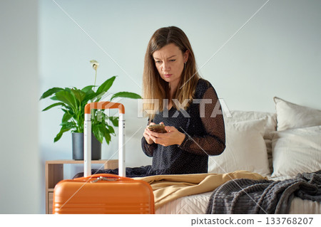 Woman in dress sitting on bed holding smartphone with suitcase nearby 133768207