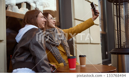Two young women taking selfie at outdoor cafe table with takeaway coffee cups Two young women taking selfie at outdoor cafe table with takeaway coffee cups 133768494