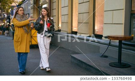 Two women walking with coffee cups in urban street conversation during autumn day Two women walking with coffee cups in urban street conversation during autumn day 133768498