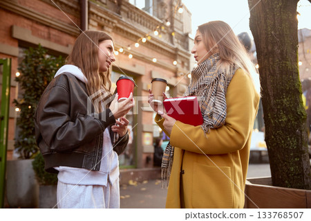 Two young women chatting outdoors with coffee cups and notebook in autumn street scene Two young women chatting outdoors with coffee cups and notebook in autumn street scene 133768507