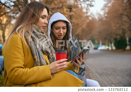 Two young women with coffee outdoors checking smartphone in autumn scenery 133768512