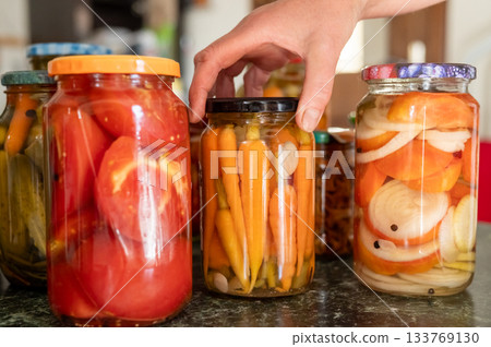 Hand reaching for pickled jars with assorted vegetables on kitchen counter. National Pickle Day 133769130