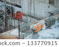 Red Rusty Hook suspension Tower Crane against background Warehouse of Yellow Volumetric I-Beams Formwork on Roof Monolithic Building Box. Background Construction Site with Rebar outlet Concrete Walls. 133770915