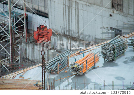 Red Rusty Hook suspension Tower Crane against background Warehouse of Yellow Volumetric I-Beams Formwork on Roof Monolithic Building Box. Background Construction Site with Rebar outlet Concrete Walls. Red Rusty Hook suspension Tower Crane against background Warehouse of Yellow Volumetric I-Beams Formwork on Roof Monolithic Building Box. Background Construction Site with Rebar outlet Concrete Walls. 133770915