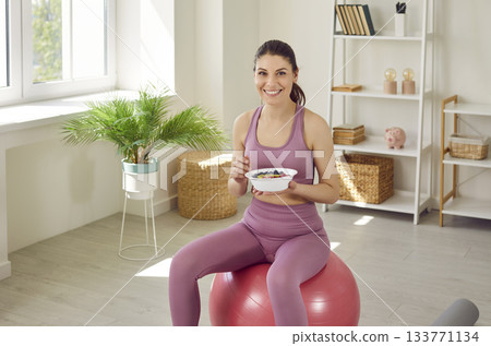 Sporty young woman sitting on fit ball and eating healthy food after working out at home. 133771134