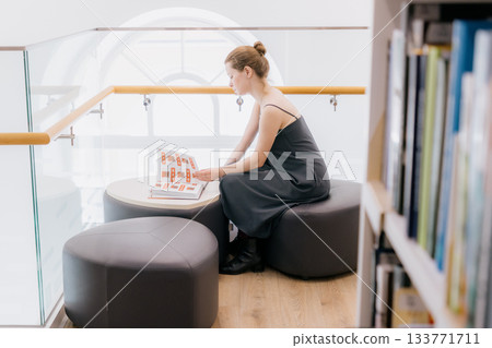 woman in library is reading book sitting on wide chair. lady in black dress flips through pages. woman in library is reading book sitting on wide chair. lady in black dress flips through pages. 133771711
