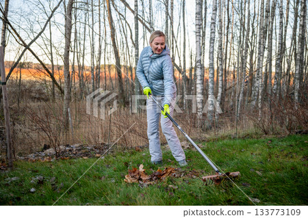 woman raking autumn leaves in forest backyard at sunset 133773109