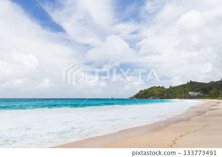 Sunlit tropical beach with clear turquoise waves, soft sand, and distant green hills 133773491