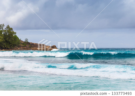 A tropical beach scene with turquoise water, white surf, and a rocky coastline 133773493