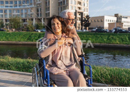 Senior woman in wheelchair walking with caregiver old man on road in park. Elderly family couple man supporting embracing woman in chair for people with disability outdoor. Rehabilitation 133774553