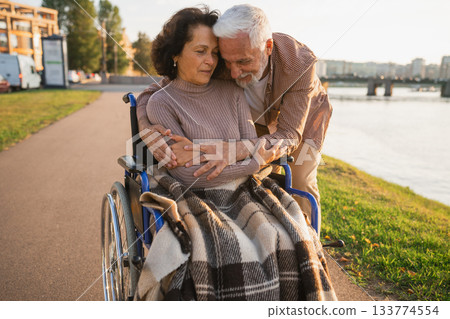 Senior woman in wheelchair walking with caregiver old man on road in park. Elderly family couple man supporting embracing woman in chair for people with disability outdoor. Rehabilitation 133774554
