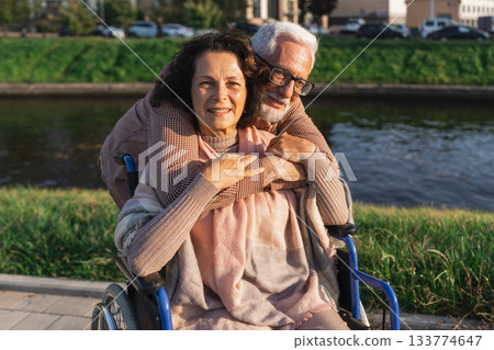 Senior woman in wheelchair walking with caregiver old man on road in park. Elderly family couple man supporting embracing woman in chair for people with disability outdoor. Rehabilitation Senior woman in wheelchair walking with caregiver old man on road in park. Elderly family couple man supporting embracing woman in chair for people with disability outdoor. Rehabilitation 133774647