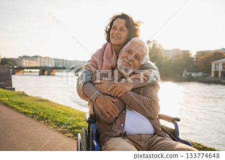 Old man in wheelchair walking with caregiver senior woman on road in park. Elderly family couple woman supporting embracing paralyzed man in chair for people with disability outdoor. Rehabilitation 133774648
