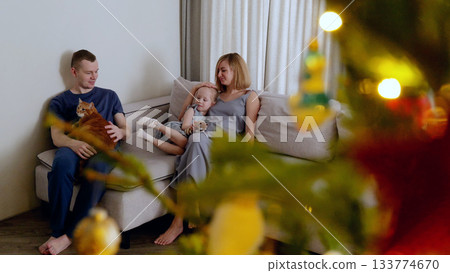 Night before Christmas. Family relaxes on the couch in their living room, basking in the cozy ambiance of the christmas tree lights and the presence of their beloved pet cat 133774670