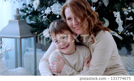 Mother and her son enjoying a heartwarming moment in front of a beautifully decorated christmas tree, surrounded by the festive spirit. A day with family before Christmas. New Year's Eve with family Mother and her son enjoying a heartwarming moment in front of a beautifully decorated christmas tree, surrounded by the festive spirit. A day with family before Christmas. New Year's Eve with family 133774675