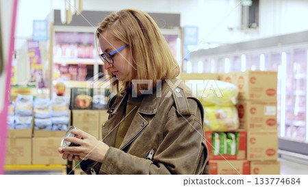 Concentrated woman shopper with glasses examining the label of a canned product in a supermarket aisle, making educated choices about her grocery items 133774684